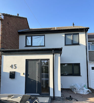 Photo of house with Garage Conversion. A Two Storey House with a Single Storey element protruding forward, with a wide anthracite front door to the right hand side of the front elevation and the door number central in the blank, off-white, rendered wall to the left of the door. Three windows are visible on the main house which is two storey.