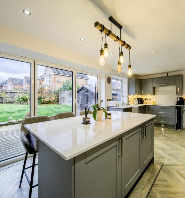 Photo of a kitchen taken at an angle, with a grey island in the centre, with white marble worktop and an industrial style light fitting above with 5 lightbulbs hanging above the island. 3 large bifold doors sit behind the island, with the rest of the kitchen visible in the back corner of the room. The floor has a light wood finish laid in a herringbone pattern.