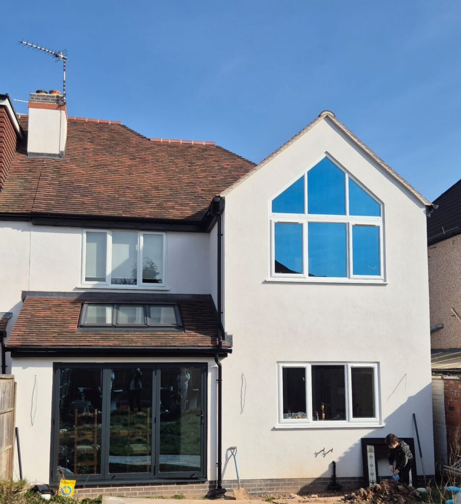 Photo of the rear of a house, with a two storey rendered gable on the right hand side, with large apex window at first floor and large window below, and a single storey extension on the left hand side with 3 panels of bifolding doors and 3 roof lights in the mono pitched roof above the doors. A standard window sits above the single storey extension.