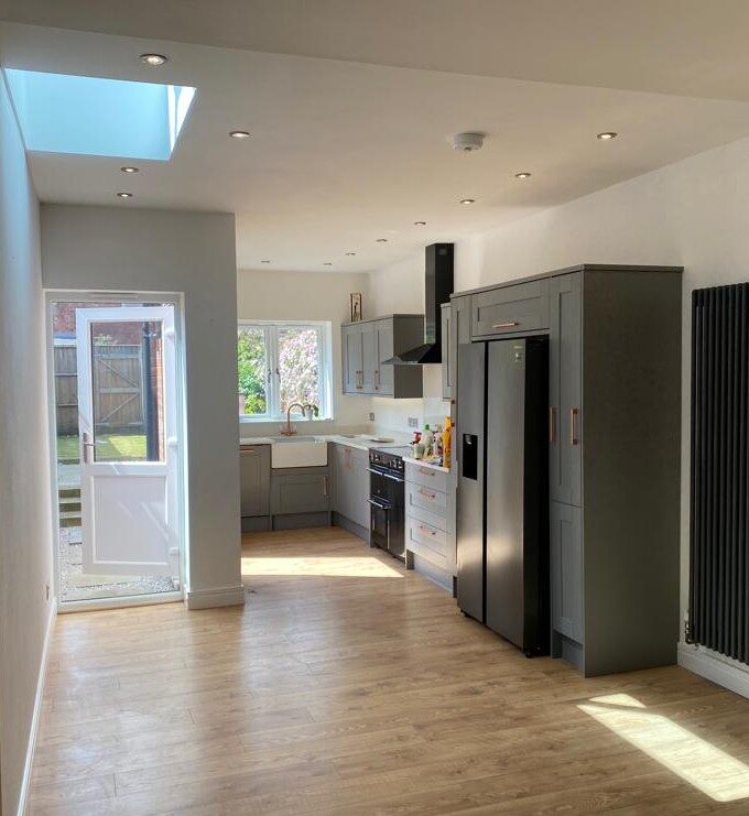 Photo of a Kitchen Extension, the photo shows a long room, with a roof lantern in the ceiling at the left of the image, with a full height window in front of it. The kitchen runs down the right hand side of the image with grey kitchen units and the kitchen window visible at the end.